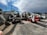 A collection of construction equipment, including multiple Bobcat machines, is parked in an outdoor lot next to a concrete wall. The scene includes excavators and loaders, all lined up on a paved surface under a partly cloudy sky.
