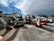 A collection of construction equipment, including multiple Bobcat machines, is parked in an outdoor lot next to a concrete wall. The scene includes excavators and loaders, all lined up on a paved surface under a partly cloudy sky.