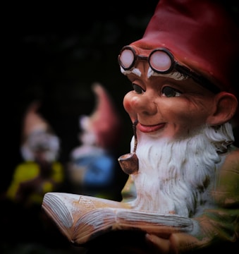 A whimsical garden gnome with a white beard and red hat is holding an open book while wearing old-fashioned glasses. The gnome appears to be smiling contentedly and has a pipe in its mouth. In the blurred background, two other gnomes can be vaguely seen, one in yellow and another in blue.