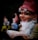 A whimsical garden gnome with a white beard and red hat is holding an open book while wearing old-fashioned glasses. The gnome appears to be smiling contentedly and has a pipe in its mouth. In the blurred background, two other gnomes can be vaguely seen, one in yellow and another in blue.
