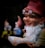 A whimsical garden gnome with a white beard and red hat is holding an open book while wearing old-fashioned glasses. The gnome appears to be smiling contentedly and has a pipe in its mouth. In the blurred background, two other gnomes can be vaguely seen, one in yellow and another in blue.