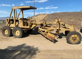 A large, yellow Caterpillar motor grader is parked on a dirt road under a clear blue sky. The machine features six large wheels, with the front-most wheel turned outward. The grader blade is positioned low to the ground, and there is a shadow cast on the ground from nearby trees or objects.