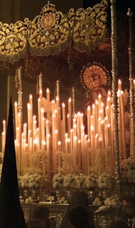 A religious procession features an ornate, gold-adorned altar with numerous lit candles. The altar includes a depiction of a religious figure with a halo. The scene is decorated with white flowers, and several shrouded figures can be seen in the foreground.