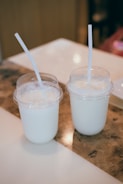 Two plastic cups with dome lids contain a creamy white beverage, each with a straw in place. They are positioned on a wooden and white surface, likely a table, with warm lighting creating a cozy atmosphere.