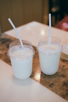 Two plastic cups with dome lids contain a creamy white beverage, each with a straw in place. They are positioned on a wooden and white surface, likely a table, with warm lighting creating a cozy atmosphere.