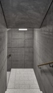 A staircase with textured concrete walls on both sides, leading to a lower level. The walls are comprised of large, rectangular concrete slabs with visible seams and bolts. Wooden handrails run along both sides of the staircase. The floor consists of light-colored, speckled tiles, and a rectangular light fixture is mounted on the ceiling, providing illumination.