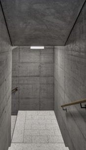 A staircase with textured concrete walls on both sides, leading to a lower level. The walls are comprised of large, rectangular concrete slabs with visible seams and bolts. Wooden handrails run along both sides of the staircase. The floor consists of light-colored, speckled tiles, and a rectangular light fixture is mounted on the ceiling, providing illumination.