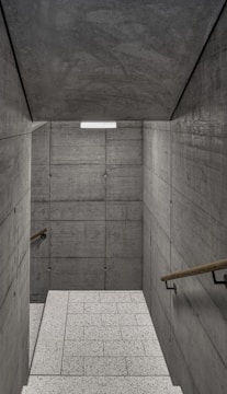 A staircase with textured concrete walls on both sides, leading to a lower level. The walls are comprised of large, rectangular concrete slabs with visible seams and bolts. Wooden handrails run along both sides of the staircase. The floor consists of light-colored, speckled tiles, and a rectangular light fixture is mounted on the ceiling, providing illumination.