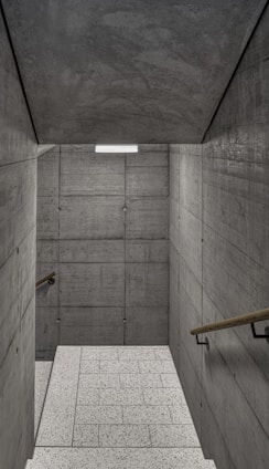 A staircase with textured concrete walls on both sides, leading to a lower level. The walls are comprised of large, rectangular concrete slabs with visible seams and bolts. Wooden handrails run along both sides of the staircase. The floor consists of light-colored, speckled tiles, and a rectangular light fixture is mounted on the ceiling, providing illumination.