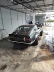 A classic sports car with a sleek design is parked in a garage-like setting. The car is a two-door coupe with wide rear tires, and the setting appears to be an industrial workshop or car service area. Two motorcycles are parked nearby, and the environment is clean with a concrete floor.