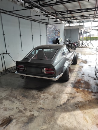 A classic sports car with a sleek design is parked in a garage-like setting. The car is a two-door coupe with wide rear tires, and the setting appears to be an industrial workshop or car service area. Two motorcycles are parked nearby, and the environment is clean with a concrete floor.