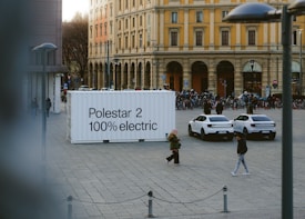 An urban square features a large white container with the text 'Polestar 2 100% electric' on it. Two white electric cars are parked nearby. People are walking around the square, and there is a row of bicycles in the background. The surrounding buildings are historic, with yellow and cream facades and large arched windows.