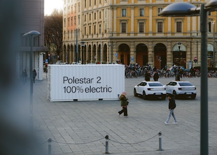 An urban square features a large white container with the text 'Polestar 2 100% electric' on it. Two white electric cars are parked nearby. People are walking around the square, and there is a row of bicycles in the background. The surrounding buildings are historic, with yellow and cream facades and large arched windows.