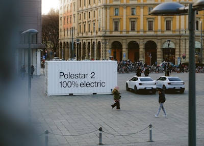 An urban square features a large white container with the text 'Polestar 2 100% electric' on it. Two white electric cars are parked nearby. People are walking around the square, and there is a row of bicycles in the background. The surrounding buildings are historic, with yellow and cream facades and large arched windows.