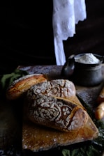 Freshly baked loaves of rustic bread rest on a wooden cutting board, intricately dusted with flour patterns. Nearby, a metal container holds a pile of flour and a wooden rolling pin is partially visible. The setting has a cozy, artisanal feel, enhanced by dark, rich textures and soft lighting.