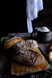 Freshly baked loaves of rustic bread rest on a wooden cutting board, intricately dusted with flour patterns. Nearby, a metal container holds a pile of flour and a wooden rolling pin is partially visible. The setting has a cozy, artisanal feel, enhanced by dark, rich textures and soft lighting.