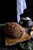 A loaf of bread with a sesame seed crust rests on a worn wooden cutting board. The background includes a rustic container filled with flour and a wooden rolling pin. A piece of white cloth hangs in the background, adding to the vintage kitchen atmosphere.