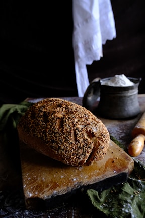 A loaf of bread with a sesame seed crust rests on a worn wooden cutting board. The background includes a rustic container filled with flour and a wooden rolling pin. A piece of white cloth hangs in the background, adding to the vintage kitchen atmosphere.