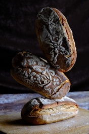 Three loaves of artisanal bread are stacked on top of each other, with intricate designs made from flour on their crusts. They are placed on a wooden surface with a dark, textured background.