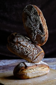 Three loaves of artisanal bread are stacked on top of each other, with intricate designs made from flour on their crusts. They are placed on a wooden surface with a dark, textured background.