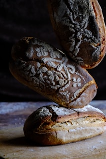 Three artisanal loaves of bread are stacked on a wooden surface. The bread features intricate flour patterns on the crust, with a rustic, hand-crafted appearance. The lighting is moody, emphasizing the texture and detail of the loaves against a dark background.