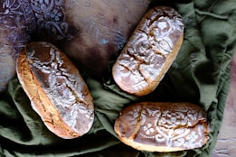 Three artisan loaves of bread are arranged on a textured, patterned surface with a green cloth beneath them. The bread has intricate dusting patterns of flour on top, highlighting the rustic and homemade quality of the loaves.