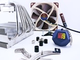 A collection of computer hardware components is arranged on a light surface. There is a large metal CPU cooler with multiple heat pipes on the left side. In the background, a brown and beige computer fan is visible, along with a tube of thermal paste. Several screws, rubber grommets, and mounting brackets are scattered around. Additionally, there is a blue keychain with the 'Core i9' logo prominently displayed.