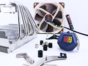 A collection of computer hardware components is arranged on a light surface. There is a large metal CPU cooler with multiple heat pipes on the left side. In the background, a brown and beige computer fan is visible, along with a tube of thermal paste. Several screws, rubber grommets, and mounting brackets are scattered around. Additionally, there is a blue keychain with the 'Core i9' logo prominently displayed.