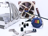 A collection of computer hardware components is arranged on a light surface. There is a large metal CPU cooler with multiple heat pipes on the left side. In the background, a brown and beige computer fan is visible, along with a tube of thermal paste. Several screws, rubber grommets, and mounting brackets are scattered around. Additionally, there is a blue keychain with the 'Core i9' logo prominently displayed.