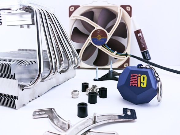 A collection of computer hardware components is arranged on a light surface. There is a large metal CPU cooler with multiple heat pipes on the left side. In the background, a brown and beige computer fan is visible, along with a tube of thermal paste. Several screws, rubber grommets, and mounting brackets are scattered around. Additionally, there is a blue keychain with the 'Core i9' logo prominently displayed.