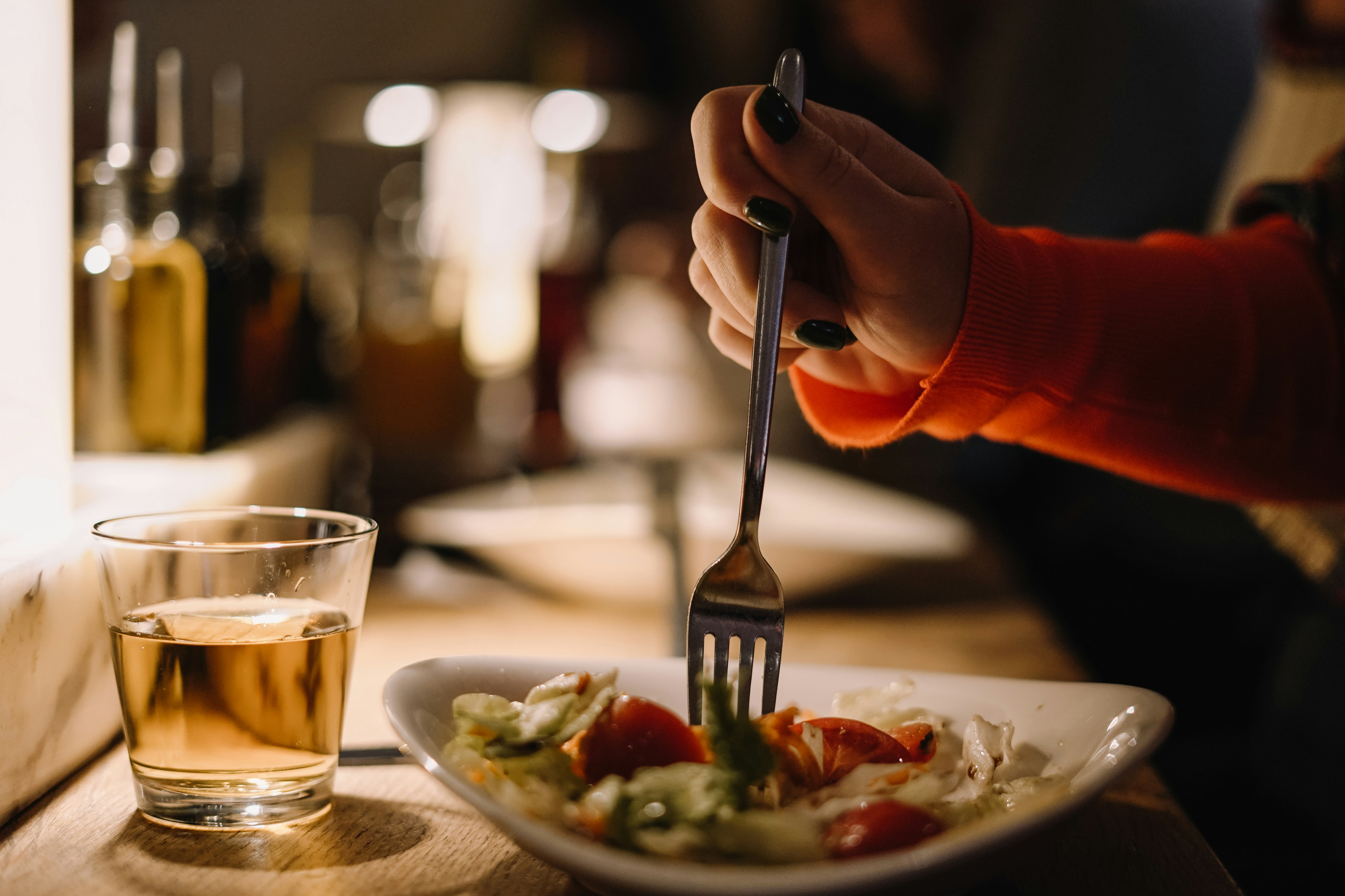 A woman in a red long sleeve shirt with black nails uses a fork to eat a salad. A cup of white wine sits next to her pla