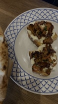 Slices of bread topped with melted cheese and saut&eacute;ed mushrooms are presented on a decorative ceramic plate with a blue and white pattern. The background shows a wooden table surface and part of a golden brown piece of bread.