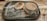 A dessert is presented in a small glass jar with a latch, topped with cocoa powder. The jar is placed on a rustic, rectangular dark plate with a dusting of powdered sugar spread across the surface. The background is a wooden table.