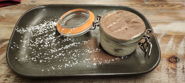 A dessert is presented in a small glass jar with a latch, topped with cocoa powder. The jar is placed on a rustic, rectangular dark plate with a dusting of powdered sugar spread across the surface. The background is a wooden table.