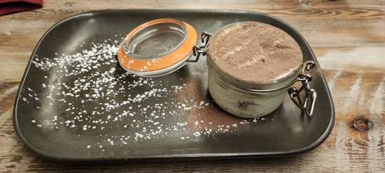 A dessert is presented in a small glass jar with a latch, topped with cocoa powder. The jar is placed on a rustic, rectangular dark plate with a dusting of powdered sugar spread across the surface. The background is a wooden table.
