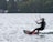 A person is engaged in kiteboarding on a lake, maneuvering skillfully across the water's surface. The individual is wearing a wetsuit and a helmet, holding onto a kite with several colorful lines. The background consists of lush trees and a few residential buildings.