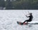 A person is engaged in kiteboarding on a lake, maneuvering skillfully across the water's surface. The individual is wearing a wetsuit and a helmet, holding onto a kite with several colorful lines. The background consists of lush trees and a few residential buildings.