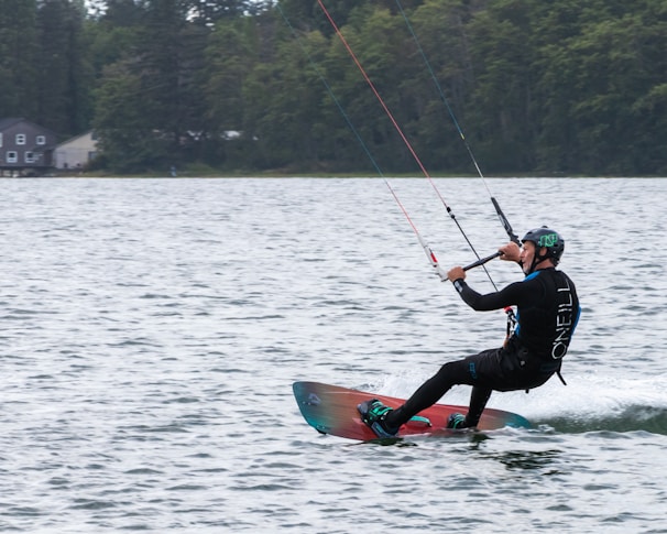A person is engaged in kiteboarding on a lake, maneuvering skillfully across the water's surface. The individual is wearing a wetsuit and a helmet, holding onto a kite with several colorful lines. The background consists of lush trees and a few residential buildings.
