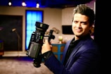 A person in a dark suit is holding a professional video camera in a room that appears to be a studio or set. The background includes a large blue illuminated window, some furniture, and various objects such as a globe.