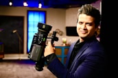A person in a dark suit is holding a professional video camera in a room that appears to be a studio or set. The background includes a large blue illuminated window, some furniture, and various objects such as a globe.