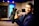 A person in a dark suit is holding a professional video camera in a room that appears to be a studio or set. The background includes a large blue illuminated window, some furniture, and various objects such as a globe.