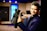 A person in a dark suit is holding a professional video camera in a room that appears to be a studio or set. The background includes a large blue illuminated window, some furniture, and various objects such as a globe.