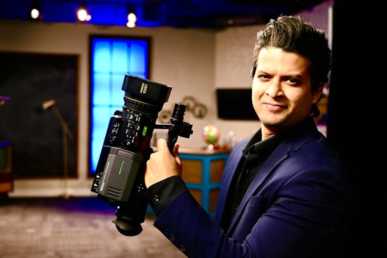 A person in a dark suit is holding a professional video camera in a room that appears to be a studio or set. The background includes a large blue illuminated window, some furniture, and various objects such as a globe.
