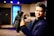 A person in a dark suit is holding a professional video camera in a room that appears to be a studio or set. The background includes a large blue illuminated window, some furniture, and various objects such as a globe.