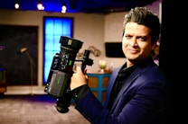 A person in a dark suit is holding a professional video camera in a room that appears to be a studio or set. The background includes a large blue illuminated window, some furniture, and various objects such as a globe.