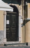 An ornate wooden door set within an arched stone frame. The door features intricate metalwork, including a knocker and a mail slot. On either side of the door are stone tiles with house numbers and a design, along with an intercom system to the right. The building wall is painted in a warm beige color with visible wear and weathering. The upper left corner shows part of an awning providing shade.