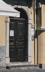 An ornate wooden door set within an arched stone frame. The door features intricate metalwork, including a knocker and a mail slot. On either side of the door are stone tiles with house numbers and a design, along with an intercom system to the right. The building wall is painted in a warm beige color with visible wear and weathering. The upper left corner shows part of an awning providing shade.