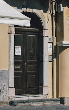 An ornate wooden door set within an arched stone frame. The door features intricate metalwork, including a knocker and a mail slot. On either side of the door are stone tiles with house numbers and a design, along with an intercom system to the right. The building wall is painted in a warm beige color with visible wear and weathering. The upper left corner shows part of an awning providing shade.