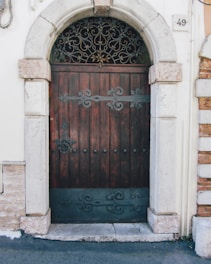 A wooden door with ornate metal decorations situated in a stone frame. The arch above the door features intricate metalwork, and there is a number 49 plaque on the wall to the right. The surrounding architecture includes white plastered walls and brick visible on one side.