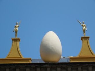 A large white egg is placed between two gold-colored statues on top of a structure. Each statue is elevated on a pedestal, and the entire setup is against a clear blue sky.